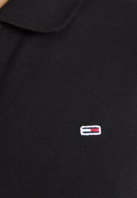 Polo noir texturé avec un petit logo rectangulaire présentant des rayures blanches, bleu marine et rouges sur la poitrine.