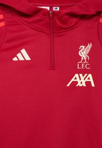 Maglia rossa con design a mezza cerniera. Presenta loghi bianchi: emblema del Liverpool FC e AXA. Include strisce bianche Adidas sulle spalle. Tessuto liscio.