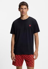 Melns kokvilnas T-krekls ar īsām piedurknēm, uz kura neliels sarkans "HUGO" logotips. Komplektā ar sarkaniem šortiem, uz kuriem atkārtoti rakstīts "HUGO".