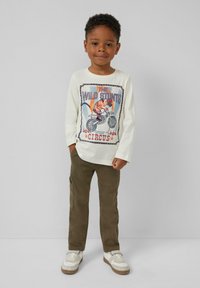 Witte langemouwen shirt met een kleurrijke grafiek van een tijger op een motorfiets, gecombineerd met olijfgroene cargo-broeken en witte sneakers.