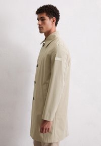 Man in een beige knooploze lange jas met een kraag, staand in profiel tegen een effen lichte muur.