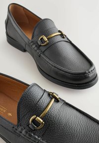 Mocasines de cuero negro con acabado texturizado, detalle de hardware en tono dorado en la parte superior y plantilla de cuero marrón. Diseño de perfil bajo.