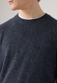 Marineblå strikkegenser laget av teksturert stoff, med spraglete mønstre og ribbestrikket krage. Lite logo-detall foran.