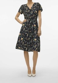 Robe à imprimé floral sur fond noir, avec manches courtes bouffantes, décolleté en V, taille cintrée et jupe évasée, portée avec des talons hauts beige.