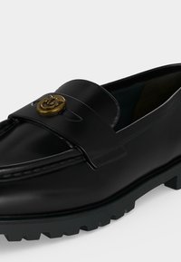 Μαύρος δερμάτινος loafer με χοντρή σόλα και χρυσή κυκλική αγκράφα στη λωρίδα μπροστά.