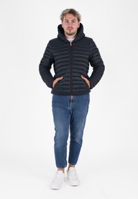 Zwarte, gewatteerde pufferjack met een capuchon en rits, met voorzakken. Gecombineerd met blauwe jeans en witte sneakers.