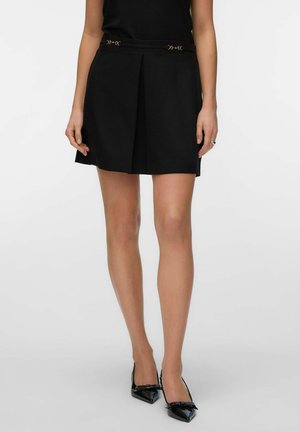 Femme portant une mini-jupe plissée noire avec des détails décoratifs en fermoirs dorés et des escarpins noirs à bout pointu avec petits nœuds.