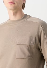 Felpa beige con collo rotondo, polsini a costine e una grande tasca sul petto con dettagli in bottone. Tessuto testurizzato con dettaglio del logo.