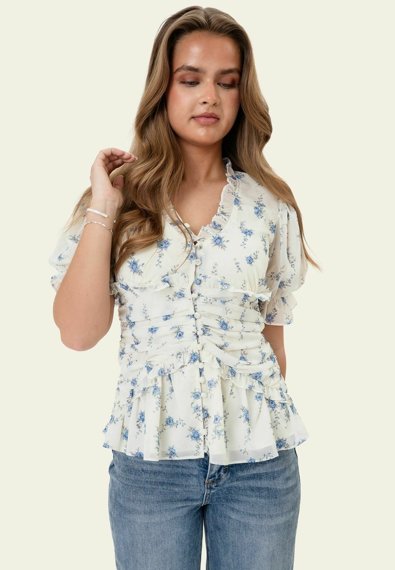 Jeune femme aux longs cheveux ondulés porte un chemisier blanc à imprimé floral bleu, avec des volants et des manches bouffantes, associé à un jean bleu, sur un fond uni.