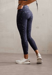 Tamsiai mėlyni ir juodi leopardų rašto leggingai su elastinga, lygia tekstūra. Dėvimi su baltais sportiniais batais, pasižyminčiais tekstūruota padu.