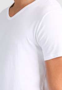 Bílý bavlněný tričko s výstřihem do V. Krátké rukávy, hladká textura, žádné vzory nebo grafika neviditelné. Jednoduchý, klasický střih.