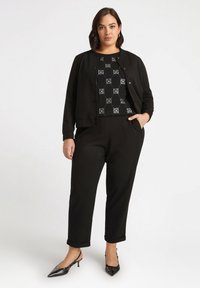 Czarna kurtka z okrągłym kołnierzem, dopasowany krój i zatrzaskami, na wzorzystą czarno-białą bluzkę, zestawiona z czarnymi, zwężającymi się spodniami i obcasami.