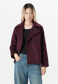 Ung kvinne med rett ombre-hår iført en mørk lilla dobbeltspent frakk, svart topp og blå jeans, som står mot en ensfarget bakgrunn.