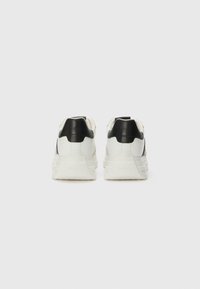 Hvite sneakers med svarte detaljer, glatt lærmateriale, polstret krage, teksturert såle og trekantet design på hælen.