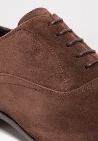 Chaussure habillée en daim marron avec une texture lisse, un bout arrondi et six lacets. Présente des coutures courbées sur le dessus et une semelle plate.