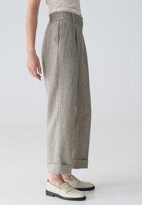 Personne portant un pantalon gris en tweed à taille haute et jambes larges avec des ourlets retroussés et des mocassins blanc cassé, debout devant un fond uni.