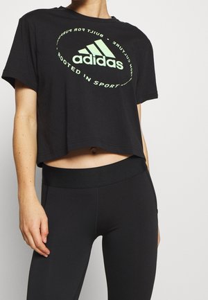 Osoba nosząca czarną krótka koszulkę Adidas z jasnozielonym logo i czarne legginsy z wysokim stanem na jednolitym tle.