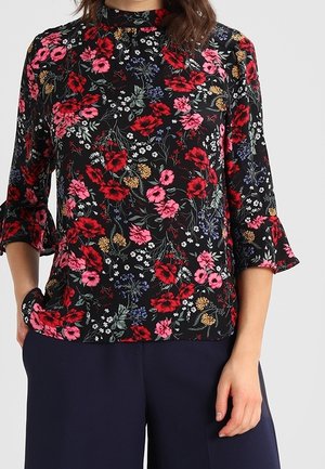Mujer que lleva una blusa negra con estampados florales en rojo, rosa y blanco y mangas de campana de tres cuartos, combinada con pantalones azul marino oscuro.