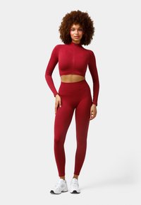 Red, fitted long-sleeve crop top with a high collar and a front zipper, paired with matching high-waisted leggings. Wearing white athletic shoes.