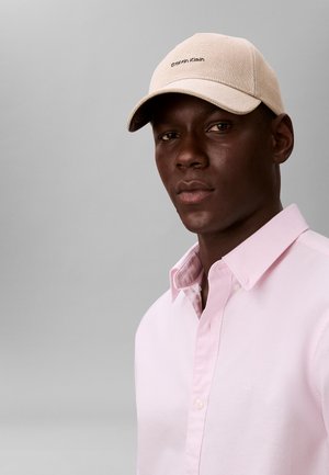 Mand iført en beige Calvin Klein-kasket og en lyserød skjorte med knapper, vendt fremad mod en ensfarvet grå baggrund.