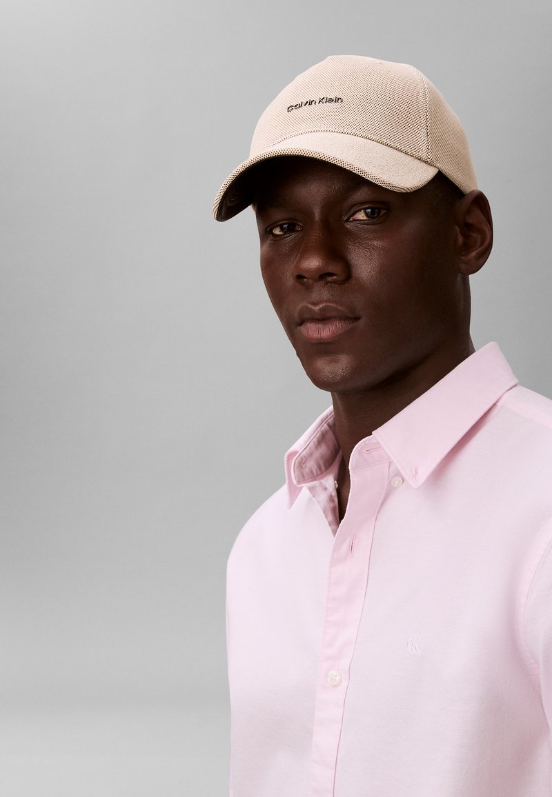 Uomo con cappellino beige Calvin Klein e camicia rosa chiaro abbottonata, davanti a uno sfondo grigio uniforme.