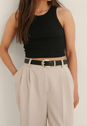 Woman wearing a black sleeveless crop top and beige high-waisted trousers with a black belt featuring a gold buckle.