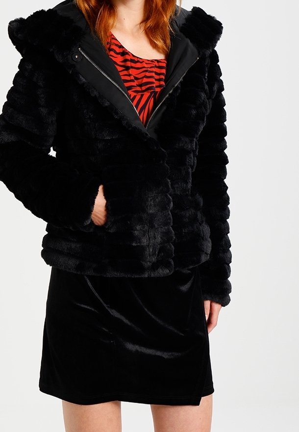 Femme portant une veste en fourrure noire texturée par-dessus un haut rayé rouge et noir et une jupe en velours noire, debout avec une main dans la poche de la veste.