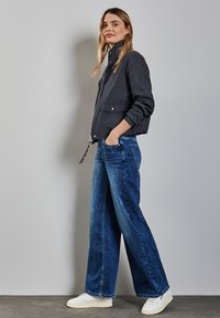 Zwarte bomberjack met een hoge kraag, gecombineerd met wijde blauwe denim jeans. Leren sneakers maken de outfit compleet. Soepele stof en casual pasvorm.