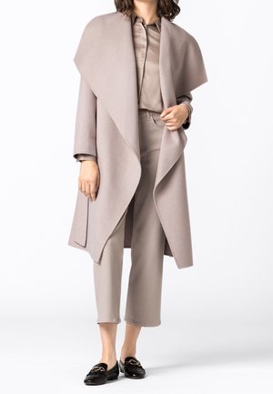 Nainen pukeutuneena taupenväriseen leveäkauluksiseen takkiin, joka on yhdistetty samansävyiseen paitaan ja lyhyisiin housuihin, sekä mustiin loafer-kenkiin, seisoo valkoista taustaa vasten.