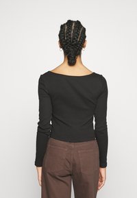 Persona con un'acconciatura a treccia raccolta, che indossa una maglietta nera a maniche lunghe e pantaloni marroni, in piedi contro uno sfondo bianco uniforme, rivolto di spalle.