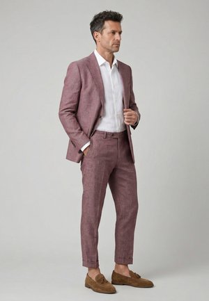 Mand iført en lavendelfarvet dragt med opsmøgede bukser, hvid skjorte og brune ruskindstasselloafers, stående mod en ensfarvet grå baggrund.