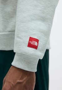 Felpa grigia chiara con polsini a coste; presenta un patch a forma di quadrato rosso con il testo "THE NORTH FACE". Il tessuto appare morbido e testurizzato.