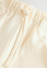 Cremefarbener Stoff mit strukturierter Oberfläche, ausgestattet mit einer geknoteten Kordel und einem elastischen Bund, was auf ein lässiges Design hindeutet.