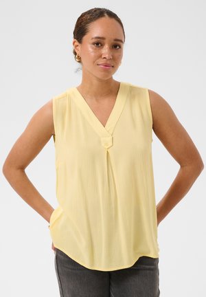 Femme aux cheveux tressés portant des boucles d'oreilles en or en forme de créoles et un chemisier jaune sans manches, debout les mains derrière le dos sur fond blanc.