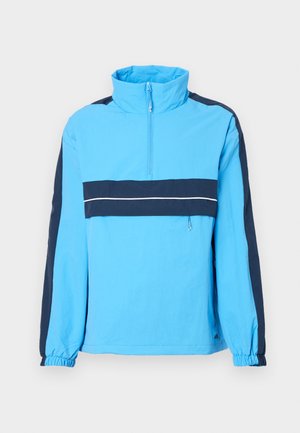 Blue pullover jacket with high collar, half-zip, navy panels, and elastic cuffs. Features a white horizontal stripe across the chest.