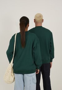 Grønne oversized hettegensere med ribbestrikkede mansjetter, båret av to personer. En har med seg en heklede kremfarget veske. Enkelt bakgrunn.
