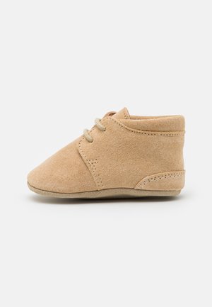 Chaussure bébé en daim doux couleur fauve avec lacets, semelle plate et coutures surpiquées, vue de profil sur fond uni.