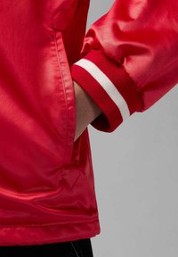 Punainen satiinitakki, jossa on punavalkoisilla raidoilla varustetut resorikahleet, sivutasku ja kiiltävä, sileä pinta.