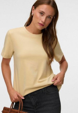 Kvinde med langt brunt hår iført en lys gul kortærmet skjorte og sorte jeans, og hun holder en brun flettet håndtaske i den ene hånd.