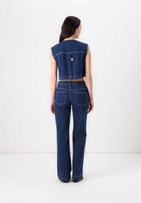 Denim prsluk s širokim ramenima i skraćenim krojem, s detaljima dugmeta na leđima. Uparen s trapericama visokog struka ravnog kroja u tamnoplavoj boji.