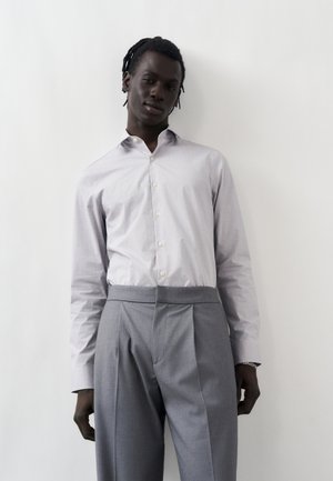 Mężczyzna w jasnej wzorzystej koszuli zapinanej na guziki, włożonej do wysokich szarych plisowanych spodni, stojący na białym tle.