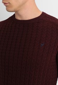 Pull en tricot torsadé bordeaux avec un col rond et un discret logo bleu sur la poitrine. La texture est en relief et douce.
