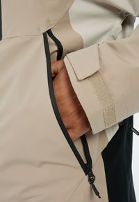 Beige en zwarte jas met een gestructureerde stof, een ritszak en een velcro sluiting bij de mouwen. De rits heeft een trekhaak voor gemakkelijke bediening.