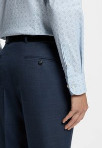 Man draagt een blauw gemêleerd overhemd met lange mouwen, ingestopt in donkerblauwe pantalon met een zwarte riem, hand rust langs het lichaam.