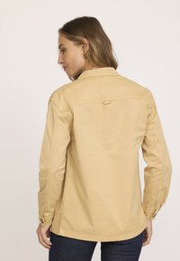 Veste en coton beige avec un col, des manches longues et des poignets boutonnés. Elle présente un empiècement arrière et des détails de couture. Texture lisse et coupe décontractée.