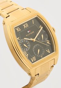 Orologio da polso in metallo color oro con quadrante nero, numeri romani, tre quadranti secondari per giorno, data e orario 24 ore, e logo "Hilfiger".