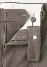Gros plan de la ceinture d'un pantalon marron montrant un bouton, une fermeture éclair, un crochet métallique et la texture du tissu.