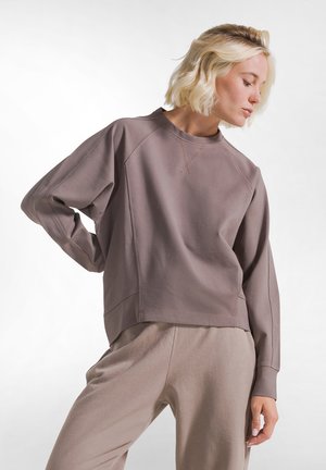 Blondynka w luźnej taupe'owej bluzie i dopasowanych spodniach, stojąca z jedną ręką na biodrze, patrząca w dół na bok.