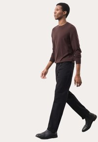 Maglione marrone a maniche lunghe, pantaloni neri e stivali neri alla caviglia. Modella che cammina, mostrando un design a vestibilità aderente e texture liscia.