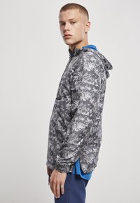 Jachetă ușoară cu glugă în model de camuflaj gri, cu față semi-zipp și manșete elastice, și tiv cu șnur. Purtați peste o cămașă albastră, vedere laterală.